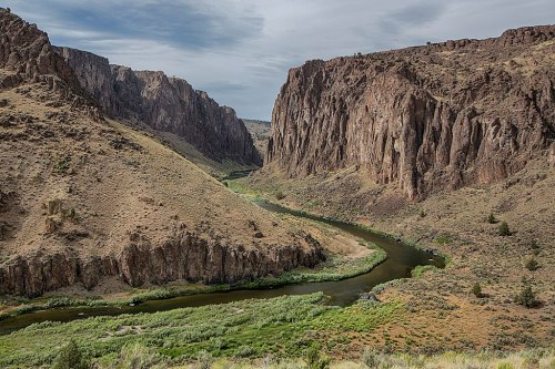 A winding river flows through a rugged canyon surrounded by steep, rocky cliffs and sparse vegetation under a cloudy sky.