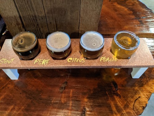 A wooden flight board with four small glasses of beer labeled Stout, PERT, Citrus, and Peach on a rustic table.