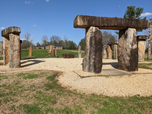 A stone structure resembling Stonehenge, surrounded by grass and trees under a clear blue sky.