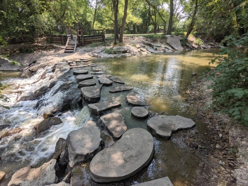 A serene creek with stepping stones, surrounded by lush greenery and trees, leading to a wooden bridge in the background.