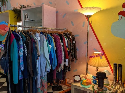 A colorful vintage clothing rack in a stylish boutique, with a lamp and decorative items against a vibrant wall.