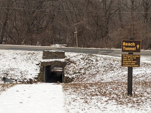 A snowy scene showing a tunnel entrance with a sign directing to the beach, near a road and wooded area.