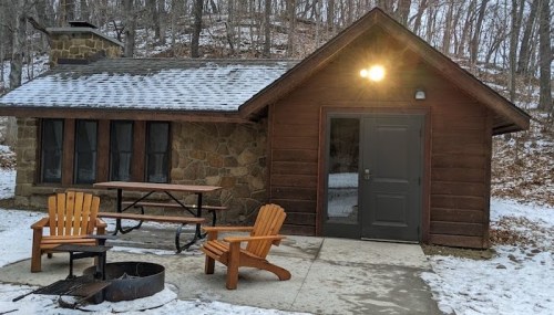 A cozy stone cabin with a wooden porch, picnic table, and fire pit, surrounded by trees and snow.