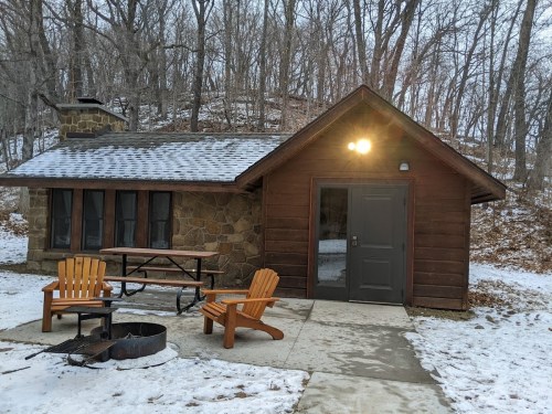 A cozy stone cabin in a snowy forest, featuring a picnic table, fire pit, and wooden chairs outside.