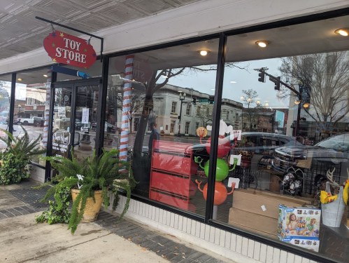 A toy store with a festive sign, displaying colorful toys and decorations in the window, surrounded by greenery.