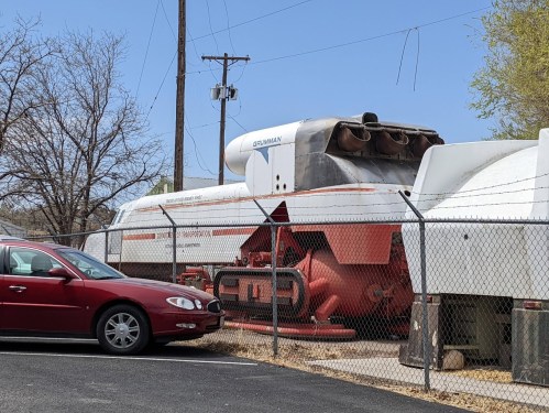 A red car parked near a large, white and orange aircraft on display behind a fence. Trees and power lines in the background.
