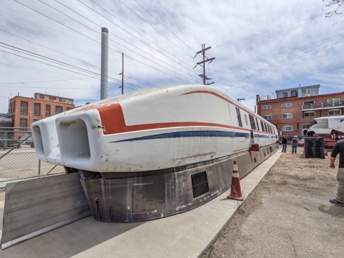 A large, futuristic train model sits on a concrete platform, surrounded by buildings and power lines under a cloudy sky.