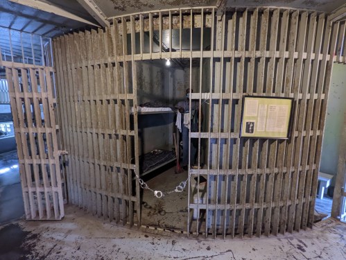 Interior of a historic prison cell with metal bars, a bed, and a display board on the wall.