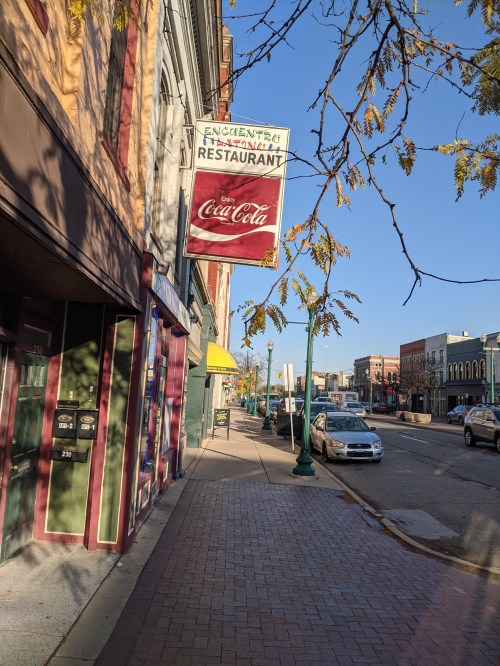 A street view featuring a restaurant sign, trees, and parked cars under a clear blue sky.