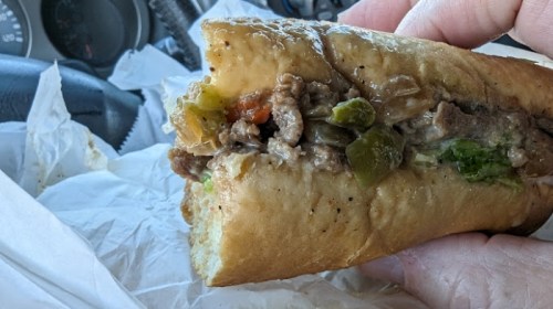A hand holding a partially eaten sandwich filled with beef, peppers, and onions, with a car interior in the background.