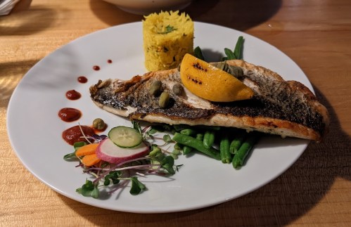 Grilled fish fillet with lemon, capers, green beans, colorful vegetables, and a serving of yellow rice on a white plate.