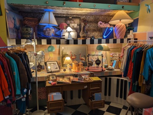 A colorful vintage shop interior with clothing racks, a piano-themed counter, and eclectic decor including lamps and flamingos.