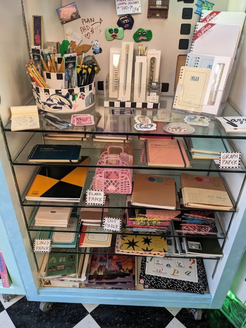 A display shelf filled with various notebooks, pens, and stationery items, organized with labels and colorful designs.