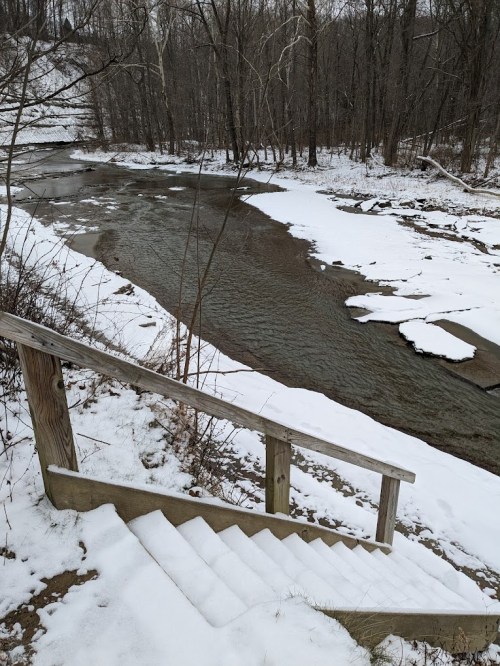 Snow-covered stairs lead down to a flowing creek, surrounded by bare trees and a winter landscape.