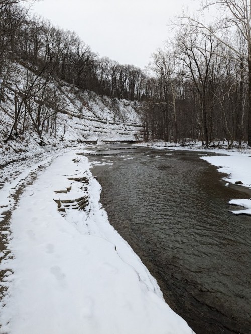 A snowy riverbank with a winding stream, surrounded by bare trees and rocky cliffs in a winter landscape.