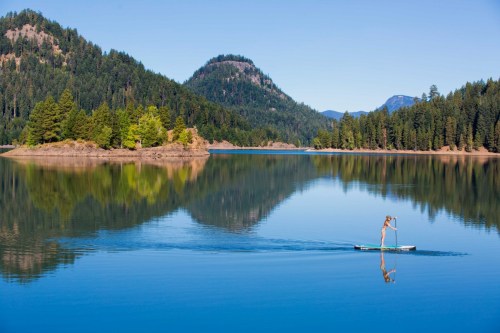 A person paddleboarding on a calm lake surrounded by lush green trees and mountains under a clear blue sky.