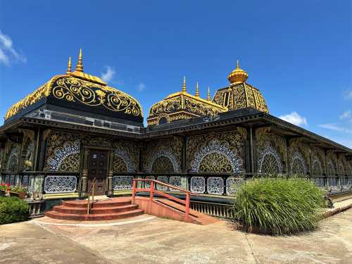 A beautifully ornate temple with golden domes and intricate designs, surrounded by greenery and a clear blue sky.