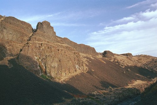 Rocky hillside with layered formations and sparse vegetation under a blue sky with scattered clouds.
