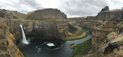 A panoramic view of a waterfall cascading into a river, surrounded by steep, rocky cliffs and lush greenery under a cloudy sky.