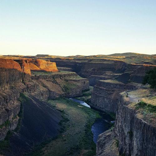A vast canyon landscape with steep cliffs and a winding river at the bottom, illuminated by warm sunlight.