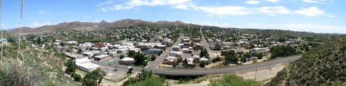 Panoramic view of a small town surrounded by mountains, featuring white buildings and a road running through the landscape.