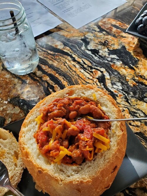 A bread bowl filled with chili and cheese, served on a textured stone table with a glass of water nearby.