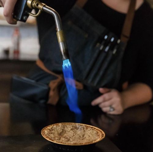 A person using a torch to caramelize the top of a dessert in a small pie dish.