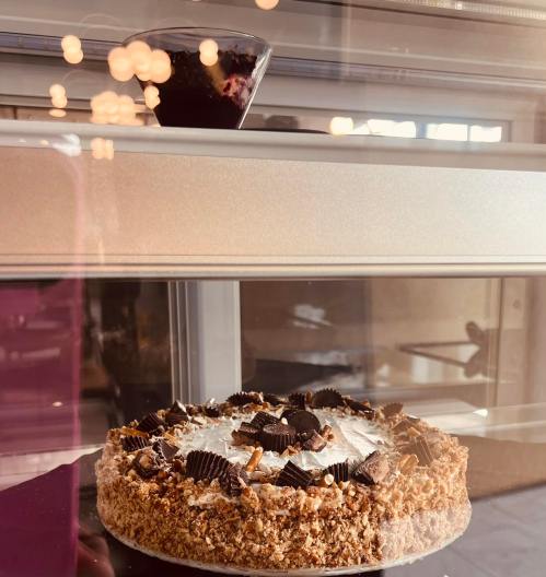 A glass display case featuring a layered cake topped with chocolate pieces and a small dessert cup in the background.