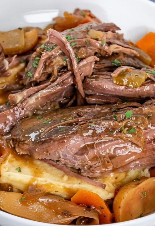 A close-up of tender beef roast served over mashed potatoes with carrots and gravy, garnished with herbs.