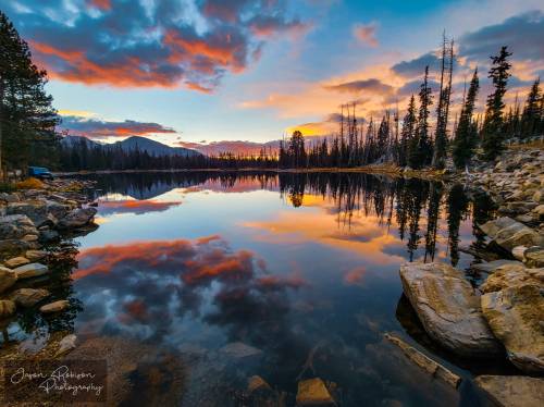 A serene lake at sunset, reflecting colorful clouds and trees, surrounded by rocky shores and a tranquil atmosphere.