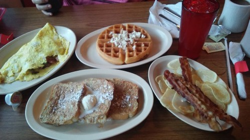 A breakfast spread featuring an omelette, waffle with toppings, French toast, and crispy bacon strips on a table.