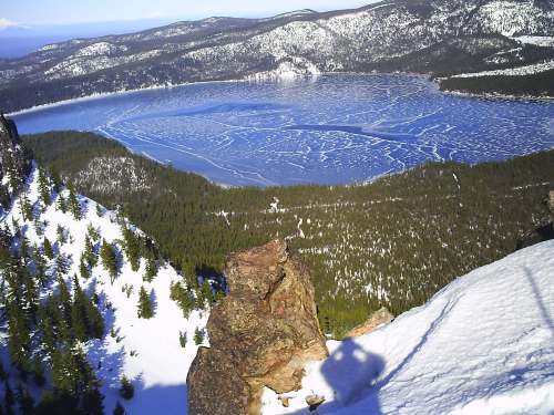 A snowy mountain peak overlooks a frozen lake surrounded by evergreen trees and distant mountains.