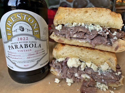 A close-up of a stacked sandwich filled with beef and blue cheese, next to a bottle of Firestone Parabola beer.