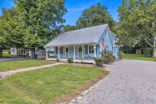 A charming blue house with a metal roof, surrounded by greenery and a gravel driveway, featuring a porch and American flag.