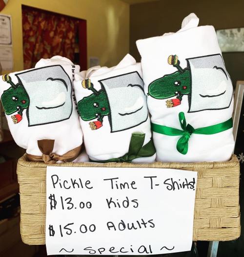 Three rolled T-shirts featuring a cartoon pickle, displayed in a basket with a sign listing prices for kids and adults.