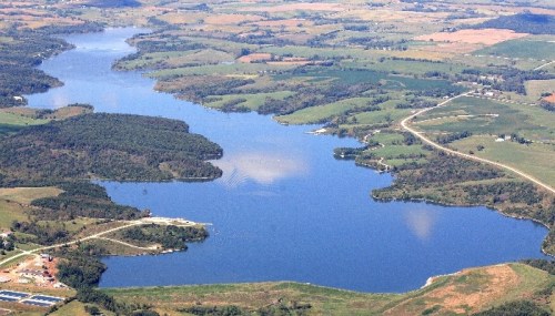Aerial view of a serene lake surrounded by lush green fields and rolling hills under a clear blue sky.