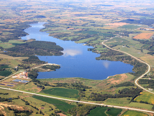 Aerial view of a large lake surrounded by green fields and patches of forest, with a winding road nearby.