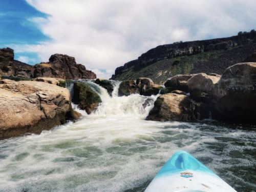 A kayak approaches a rocky waterfall, with rushing water and a scenic landscape under a cloudy sky.