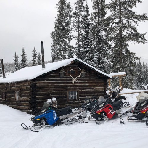 A rustic log cabin surrounded by snow-covered trees, with several snowmobiles parked in front.