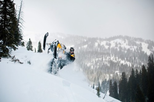 A snowmobiler performs a stunt in deep snow, surrounded by a mountainous winter landscape and evergreen trees.