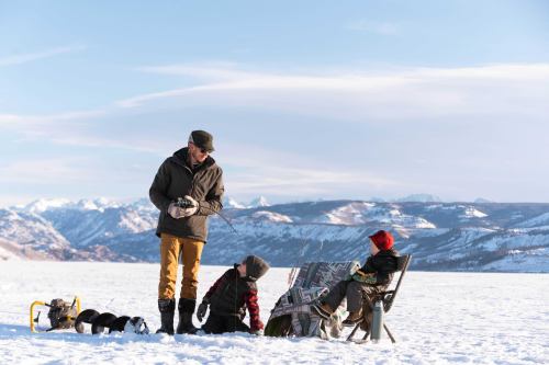 A man stands on a snowy landscape while two children sit nearby, one reading a newspaper, with mountains in the background.