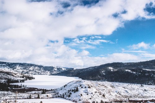 A snowy landscape with a lake, mountains, and a blue sky dotted with clouds.