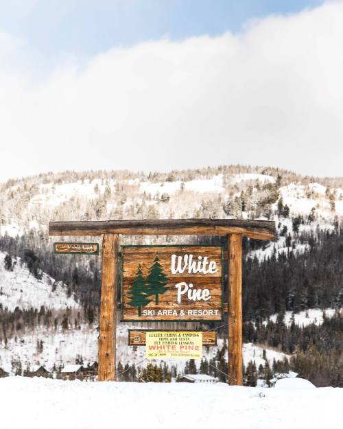 Sign for White Pine Ski Area & Resort, surrounded by snow-covered mountains and trees under a cloudy sky.