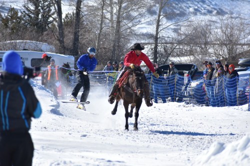 A cowboy on horseback races alongside a snowboarder in a snowy landscape, with spectators watching from the sidelines.