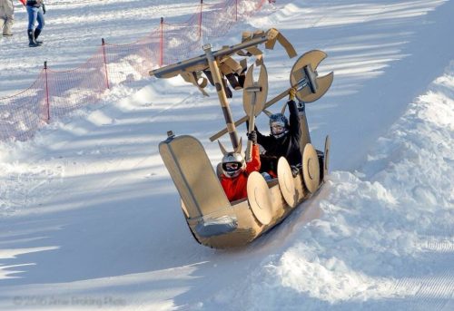 Two children in a cardboard sled shaped like a ship, joyfully sliding down a snowy hill.