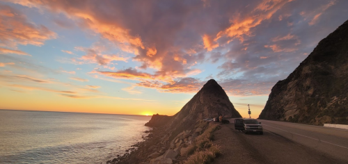 A scenic sunset over the ocean, with colorful clouds and a rocky coastline alongside a road.