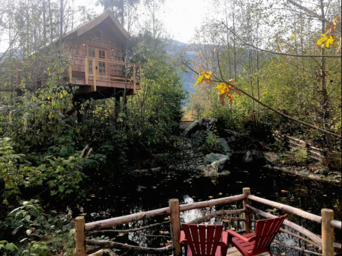 A cozy treehouse perched above a pond, surrounded by lush greenery and autumn leaves, with red chairs in the foreground.