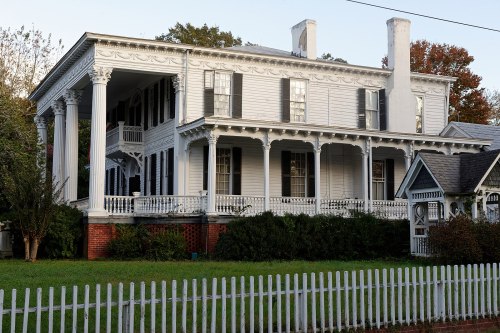 A large, historic white house with columns, black shutters, and a picket fence, surrounded by greenery.