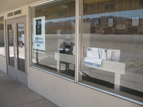 A storefront with large windows displaying a passport application sign and a table inside. Reflections of the surroundings are visible.