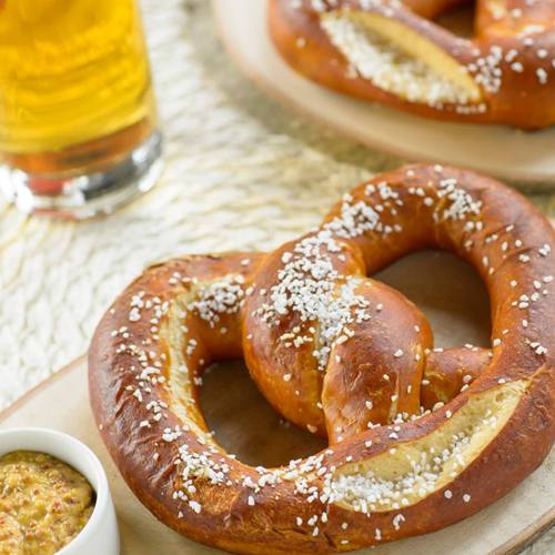 A close-up of a soft pretzel sprinkled with salt, served with a small bowl of mustard and a glass of beer in the background.